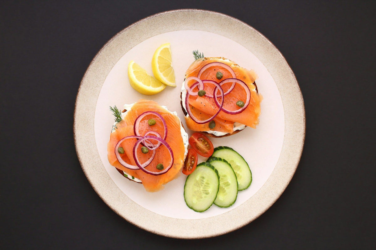 Dwie tosty z wędzonym łososiem, czerwoną cebulą i kaparami na talerzu, obok ogórek i cytryna – źródła białka i omega-3 w diecie wspierającej włosy